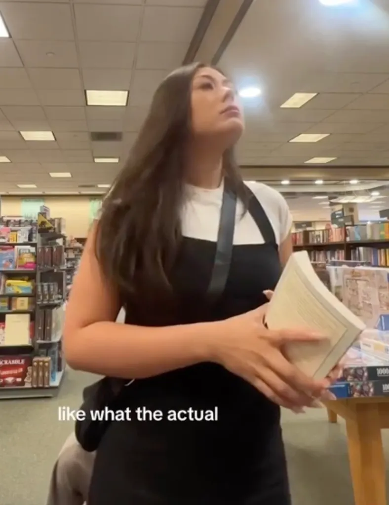 A woman in a black and white top holding a book in a bookstore, looking upwards in distress.