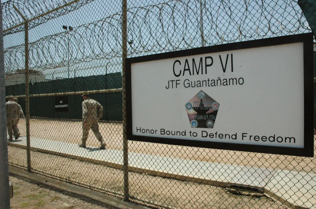 Sign marking an entrance to Camp VI detention center at Guantanamo Bay with two guards walking behind a chain-link fence.