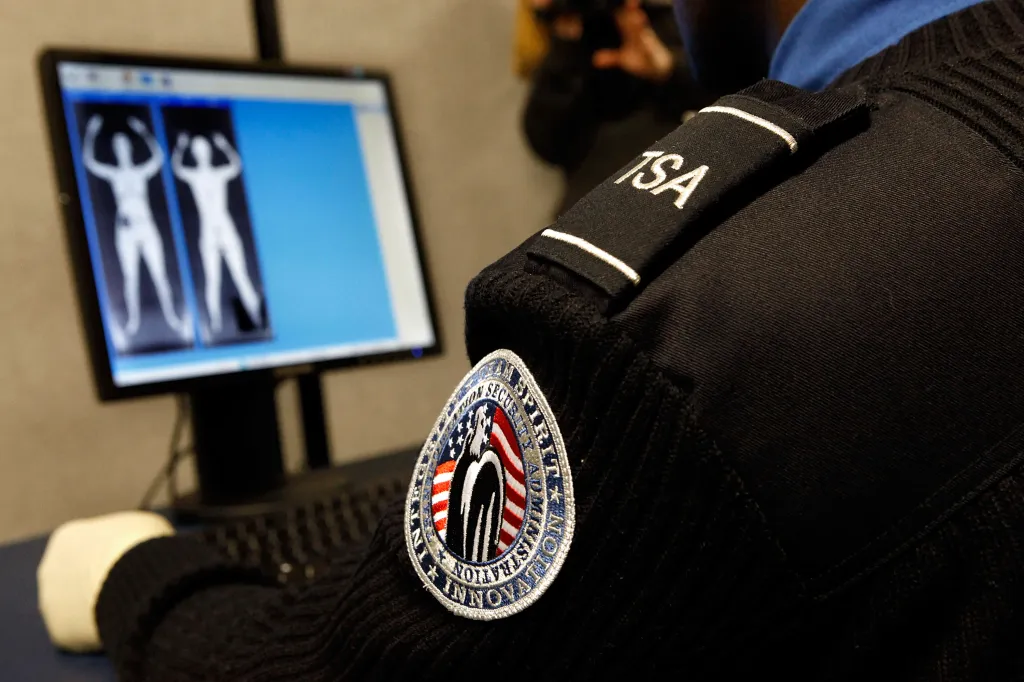 A close-up of a TSA officer's uniform shoulder and sleeve, with a computer monitor displaying backscatter scan images in the background.