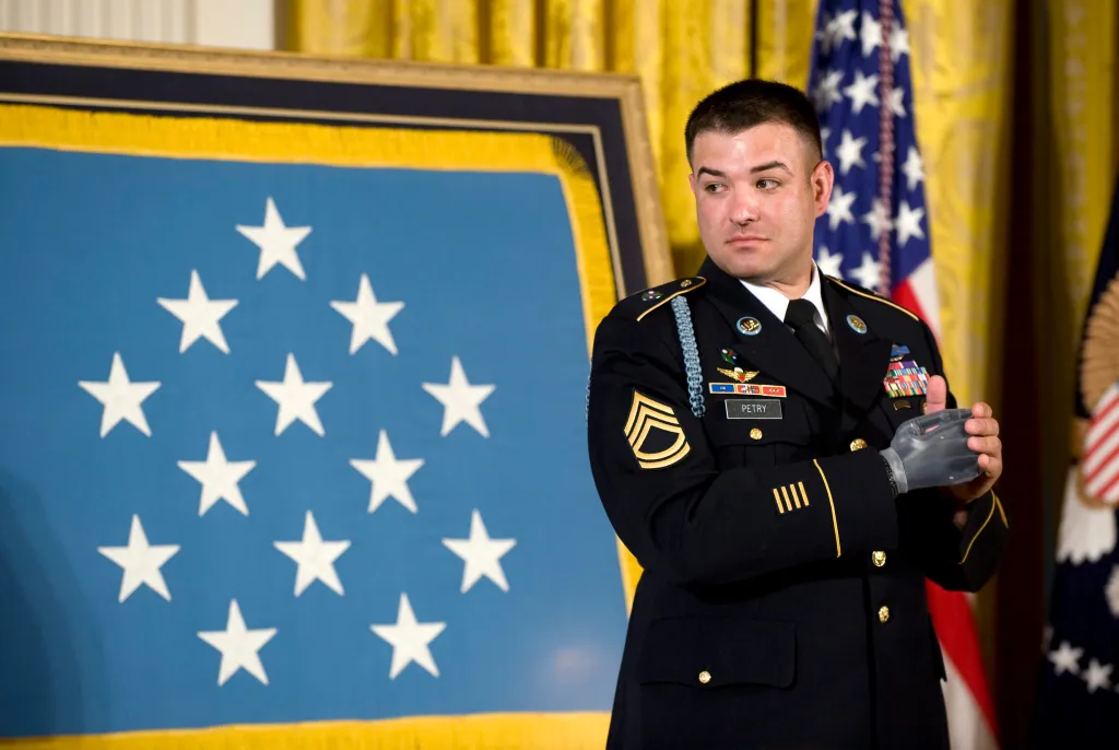 Army Sergeant First Class Leroy Arthur Petry applauding, wearing his dress uniform, with a prosthetic right hand, standing in front of a blue banner with white stars.