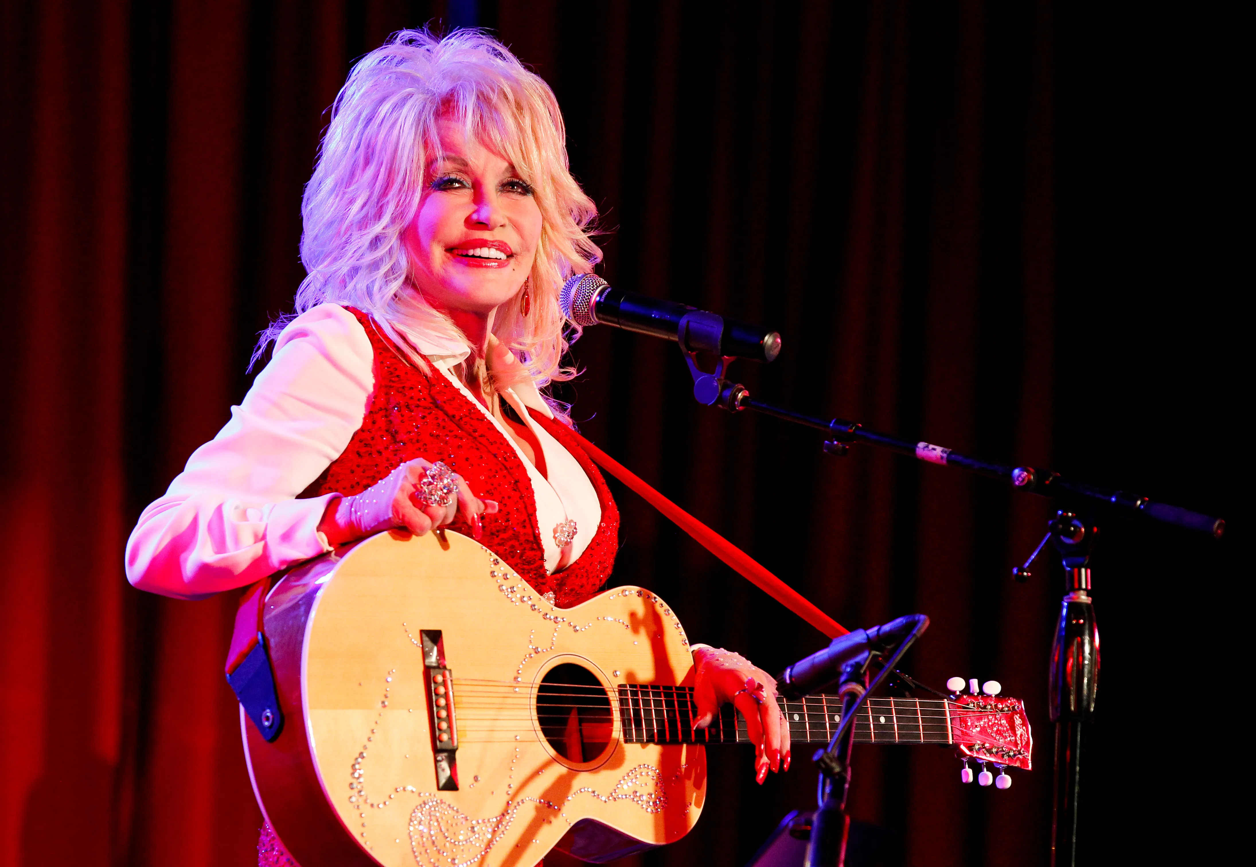 Dolly Parton holding a rhinestoned guitar while singing into a microphone.