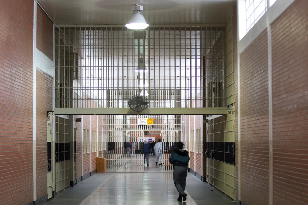 A person walks through a long prison hallway with cell blocks on either side.