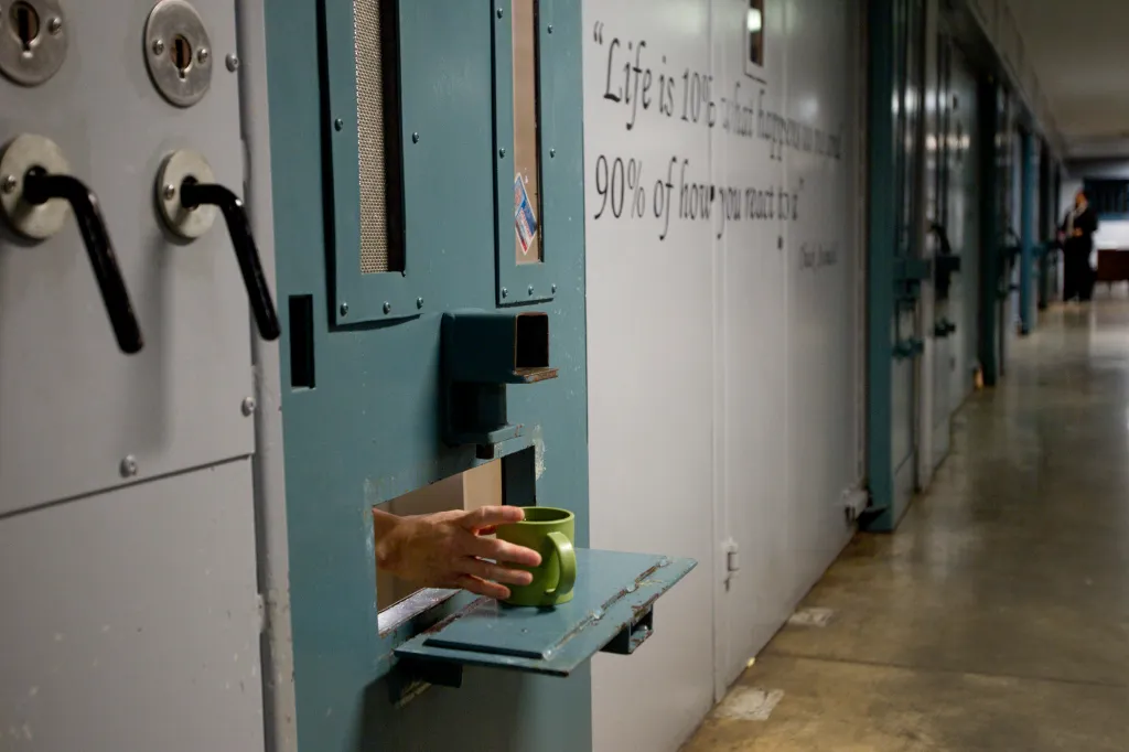 An inmate's hand reaching for a green cup through a slot in a teal prison cell door.