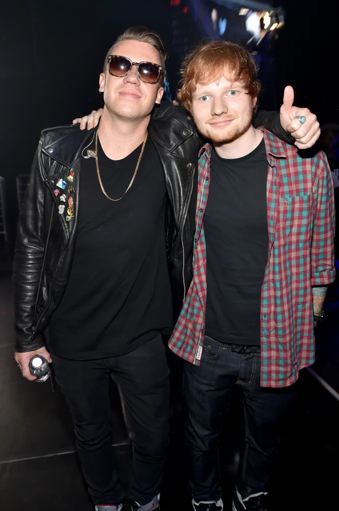 Macklemore and Ed Sheeran pose for a photo with Macklemore wearing a black leather jacket and sunglasses, and Ed Sheeran in a plaid shirt giving a thumbs-up.