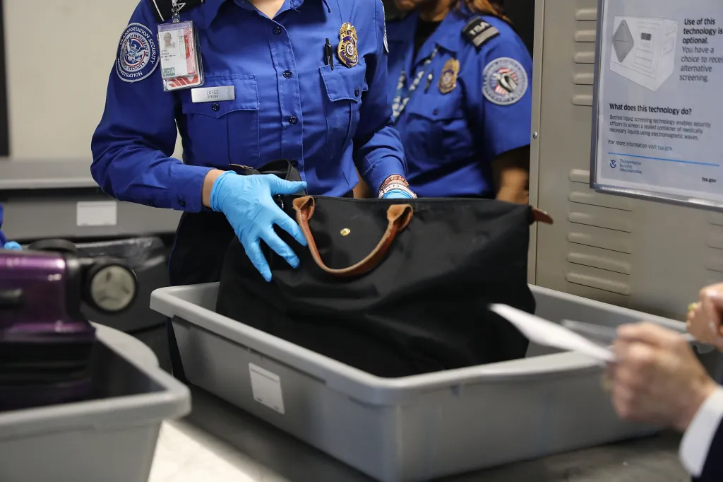 A TSA officer in blue uniform and gloves places a black bag into a gray security bin at LaGuardia Airport.