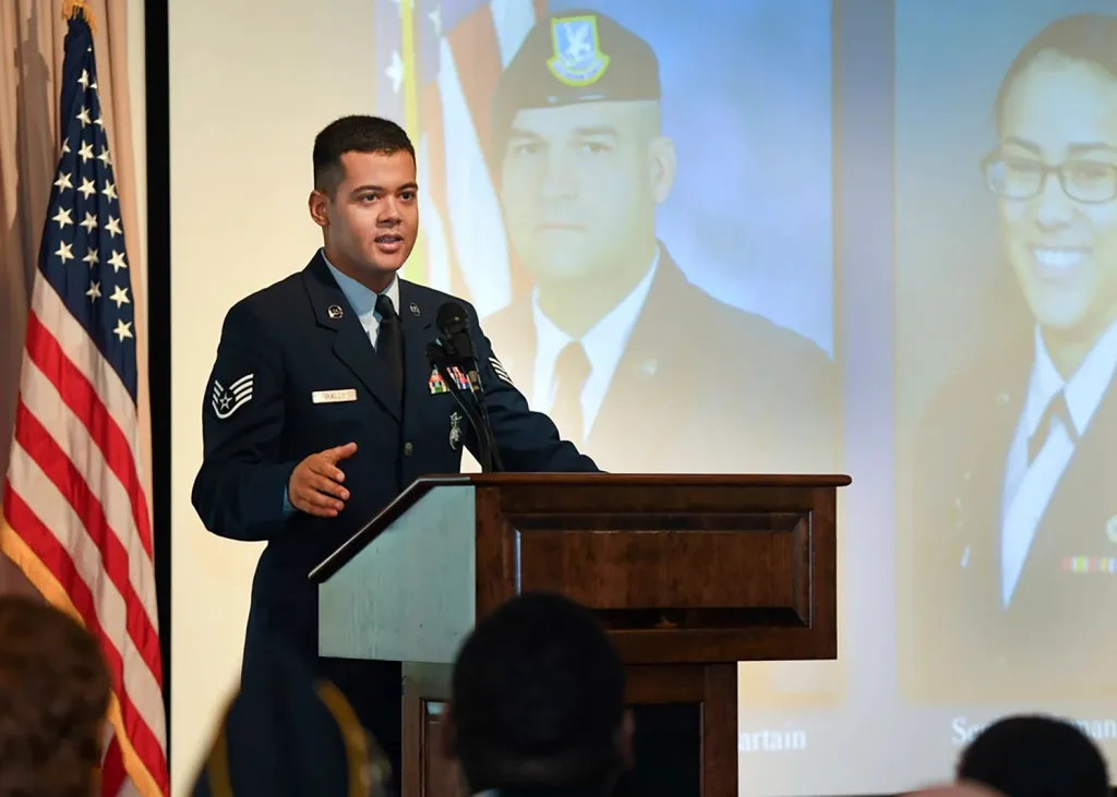 Wilmer Puello-Mota speaking at a podium with an American flag to his left and two images of people in uniform projected behind him.
