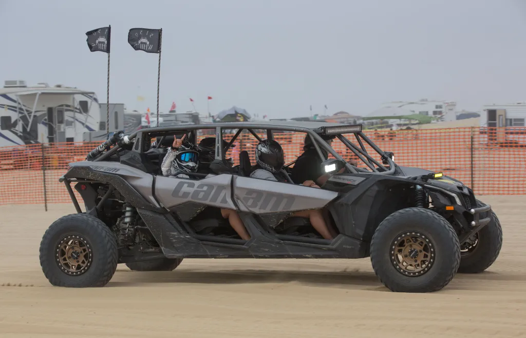 An off-road vehicle with four people in it drives on a sandy beach.