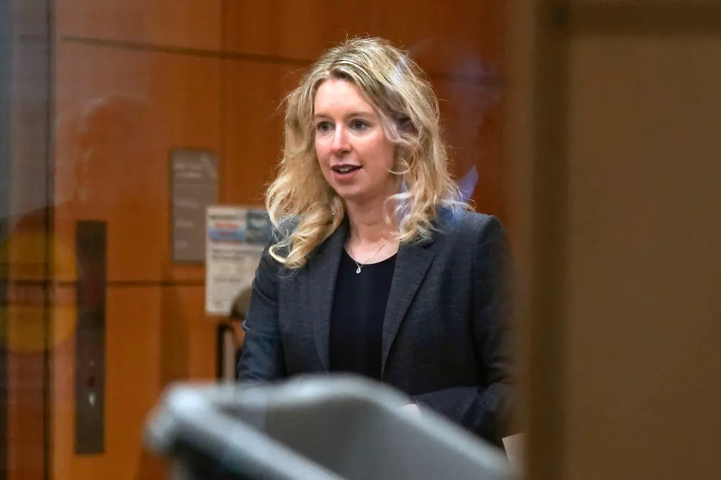 Elizabeth Holmes, former Theranos CEO, in a black shirt and gray blazer, looking forward with her mouth slightly open.