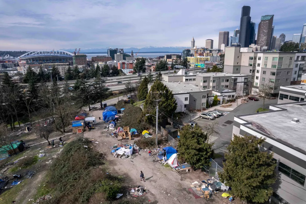 An aerial view of a homeless encampment covered in garbage, with downtown Seattle and the Seattle skyline in the background.