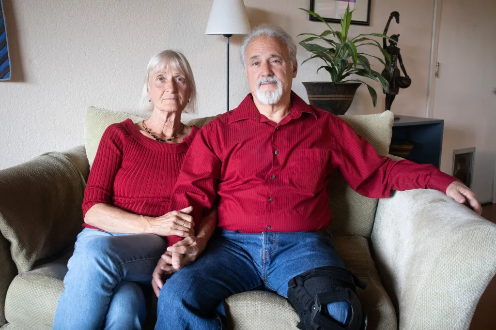 Jeff and Els Woodke, wearing red shirts and blue jeans, sitting on a couch.