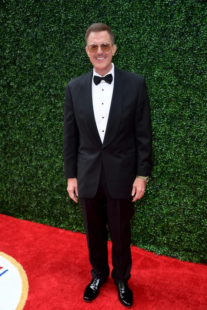 Billy Gardell poses in a black tuxedo and sunglasses on a red carpet.