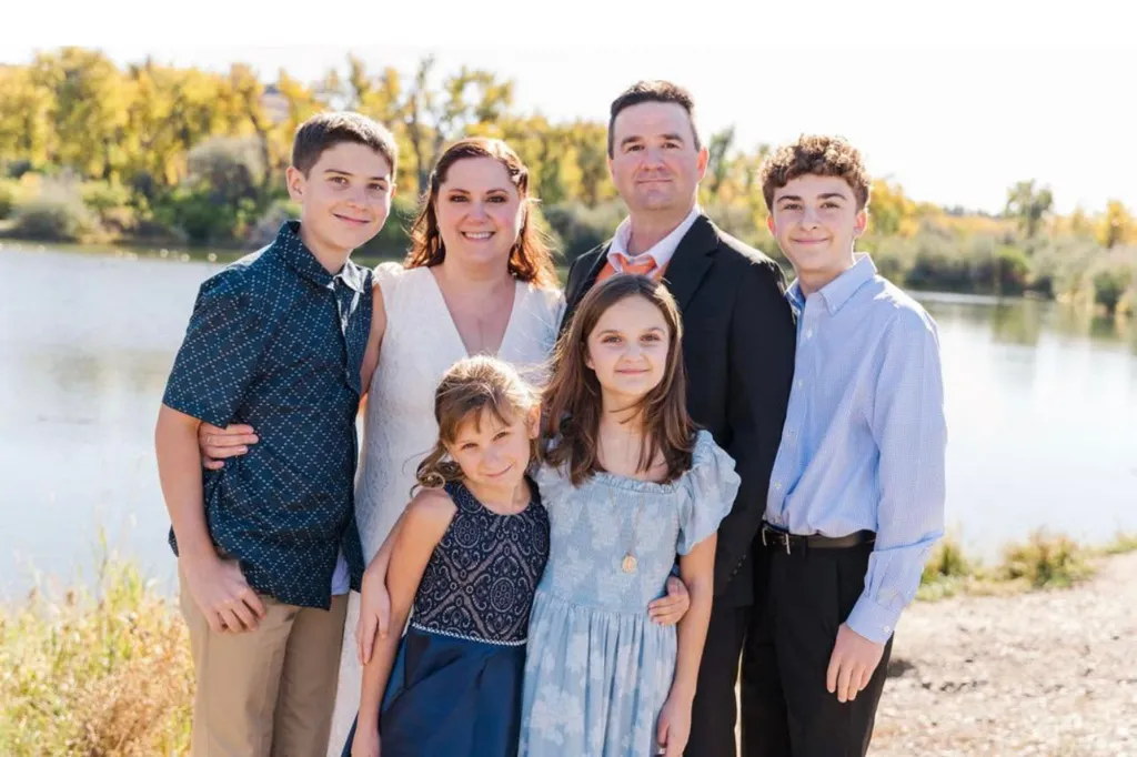 The Ghekiere-Ireland family, including Owen Ireland, Megan Ghekiere, Chloe Ireland, Charlotte Ghekiere, Adam Ghekiere, and Landon Ghekiere, stands by a body of water with trees in the background.