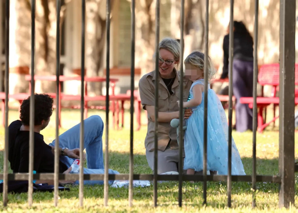 Elizabeth Holmes smiling and kneeling to play with her child in a blue dress, while her husband sits on the grass nearby.