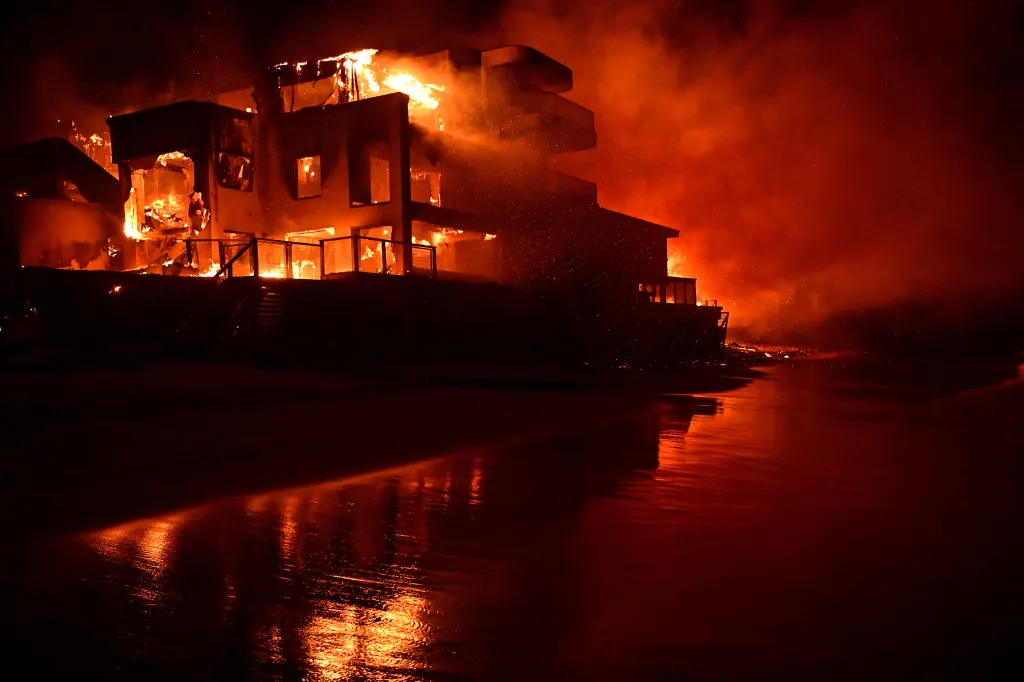 A large house on the beach engulfed in bright orange flames at night.