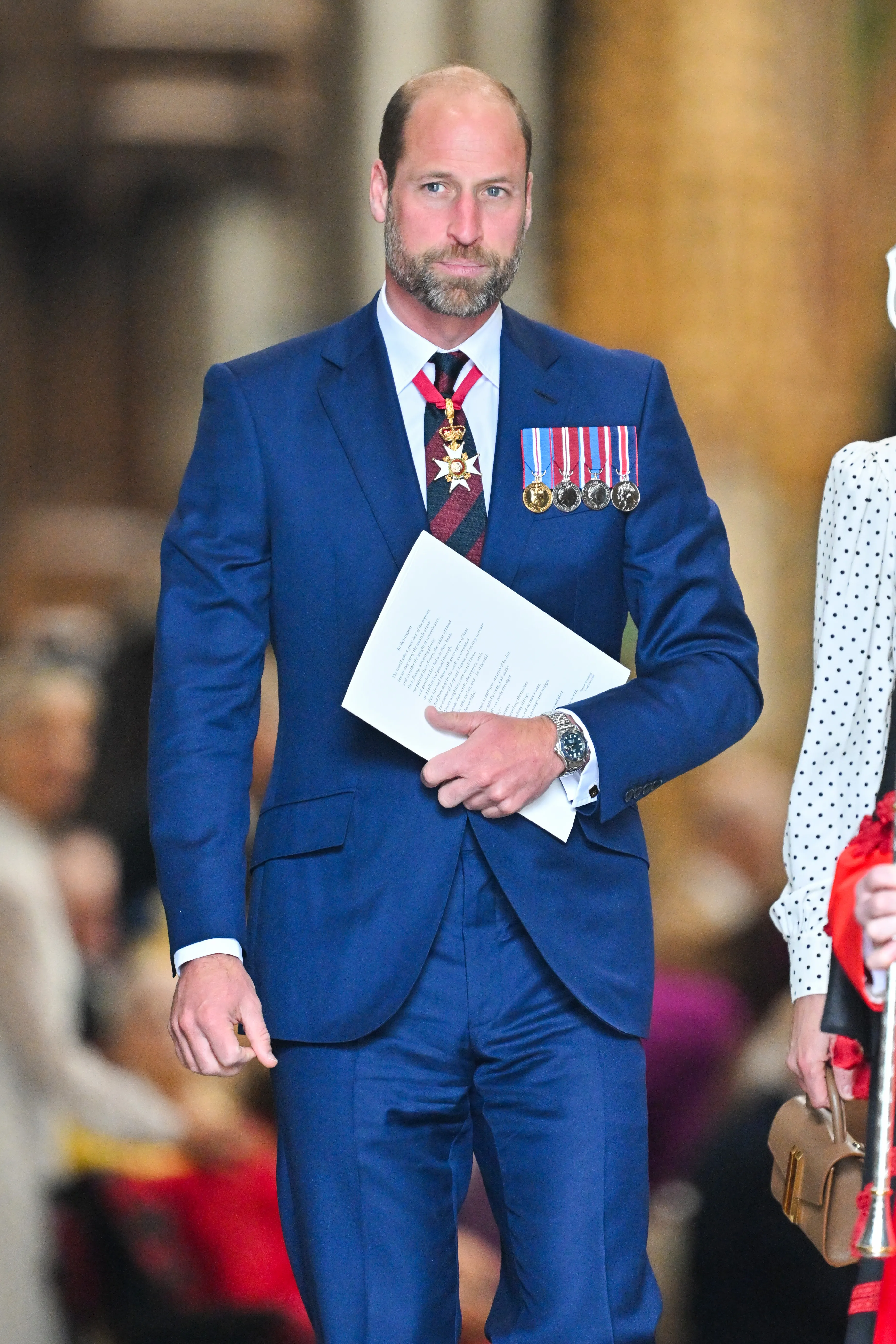 Prince William departing a thanksgiving service in a blue suit with military medals, holding a program in his hand.