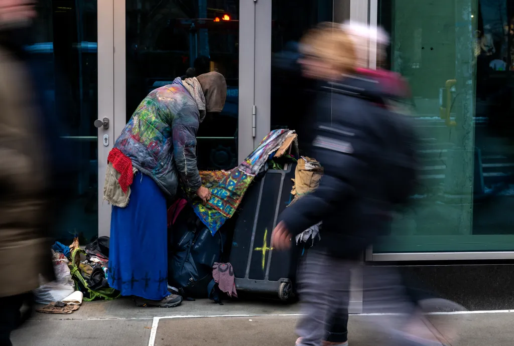 A homeless person organizing their belongings on a New York City street, with passersby blurred in motion.