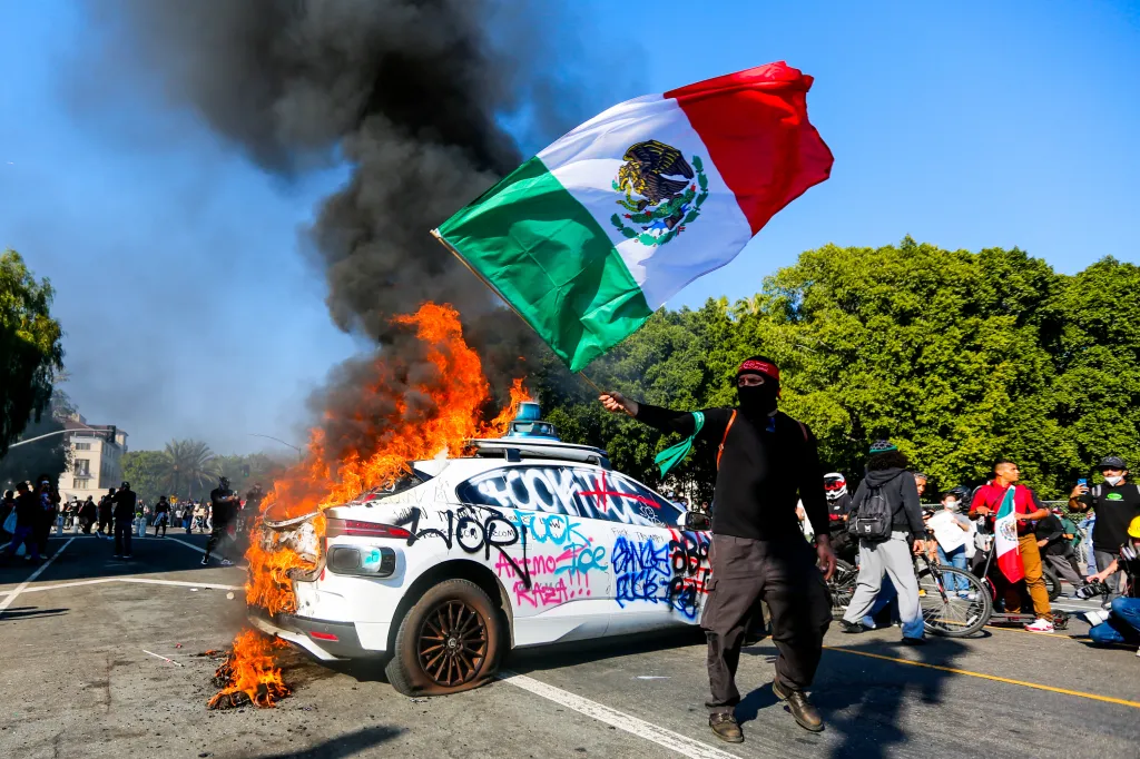 A protester waves the Mexican flag in front of a burning, graffiti-covered vehicle during an anti-ICE protest in Los Angeles.