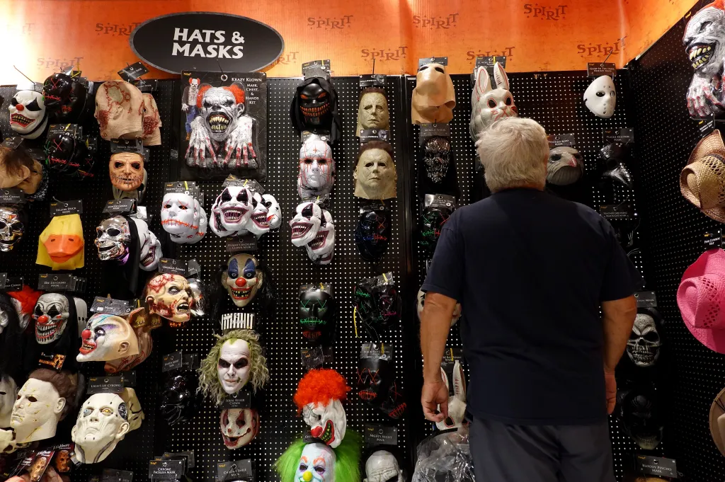 A customer shops for Halloween masks at a Spirit Halloween store, with various scary and clown masks displayed on black pegboards.