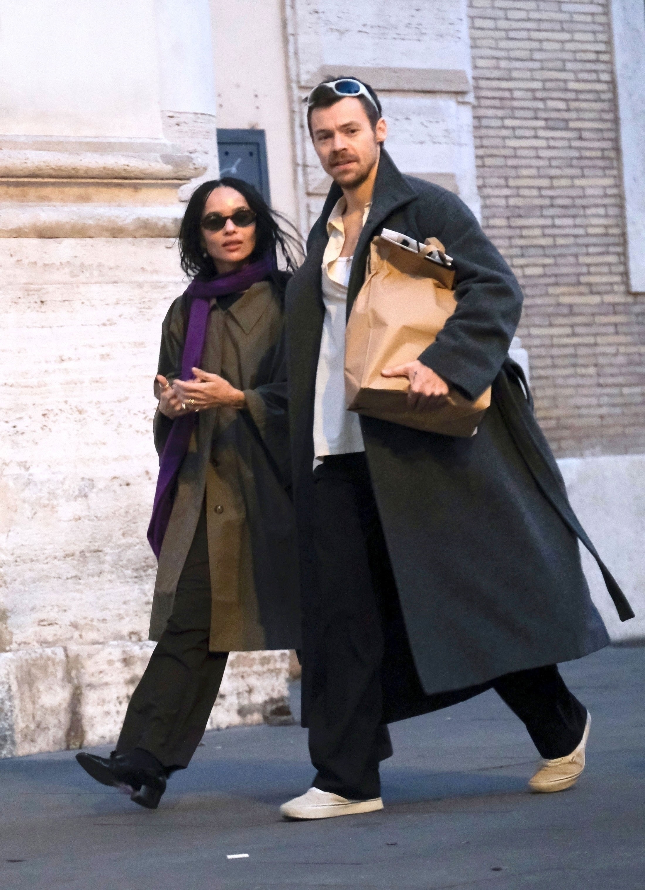 Harry Styles and Zoe Kravitz walking arm in arm on a cobbled street, Styles holding a paper shopping bag.