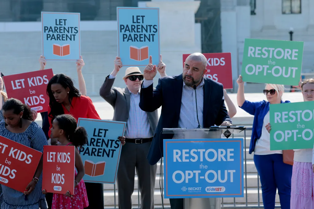 Wael Elkoshairi speaks at a podium outdoors, surrounded by supporters holding signs that read 