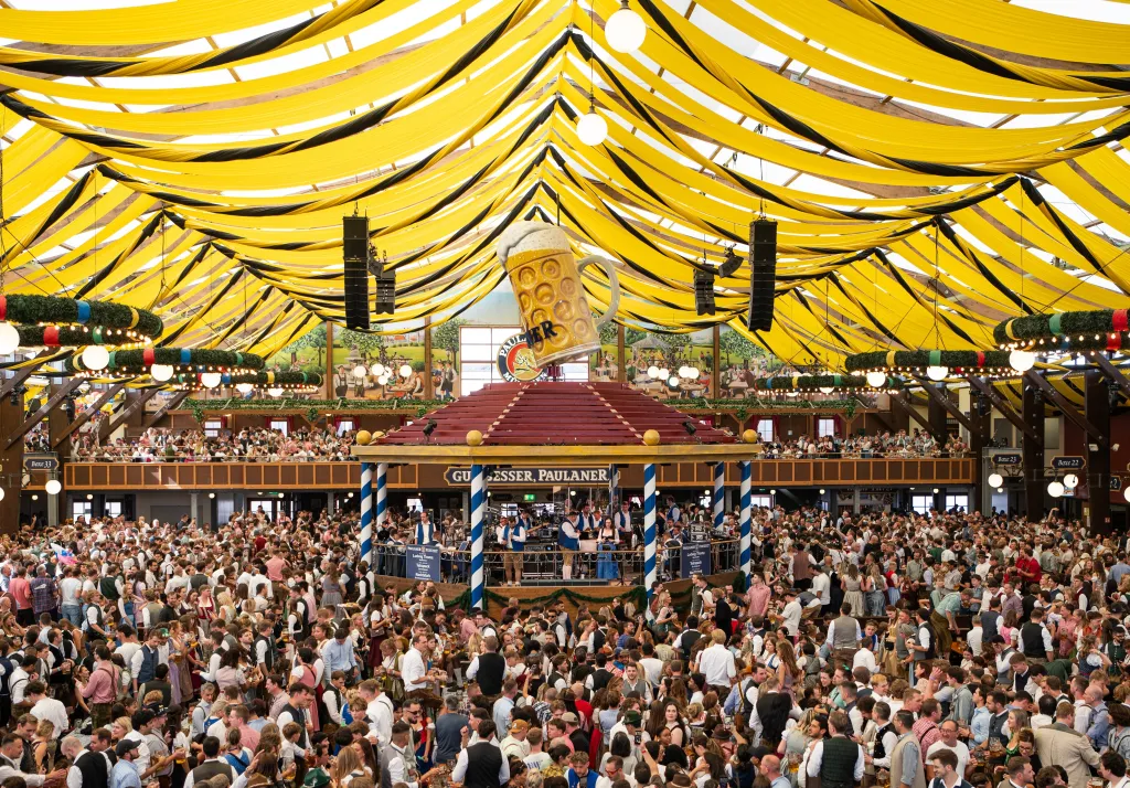 Crowds of people inside a large tent with yellow and black striped fabric draped overhead, celebrating Oktoberfest.