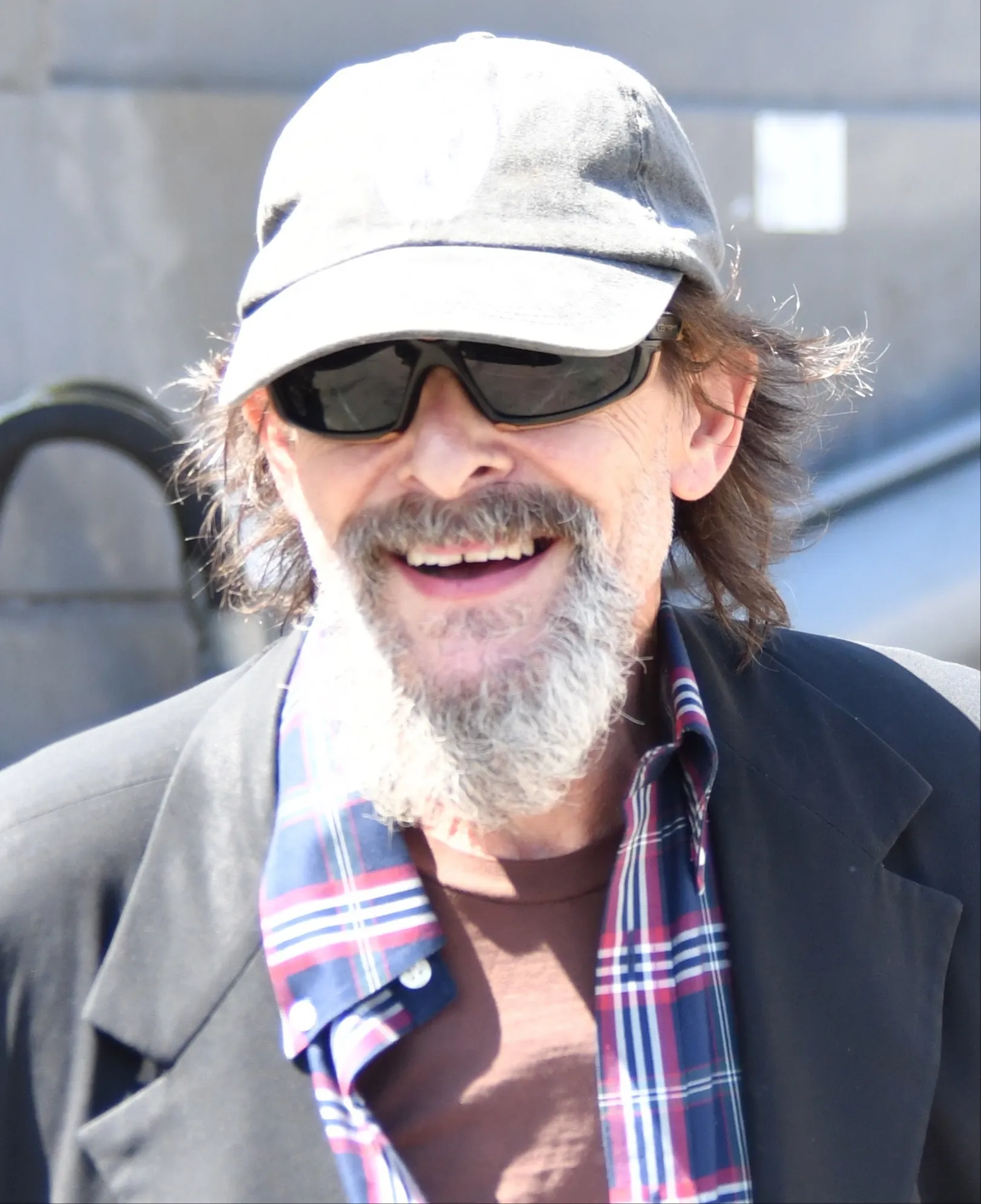 Judd Nelson smiling with a white beard, sunglasses, and a grey cap.