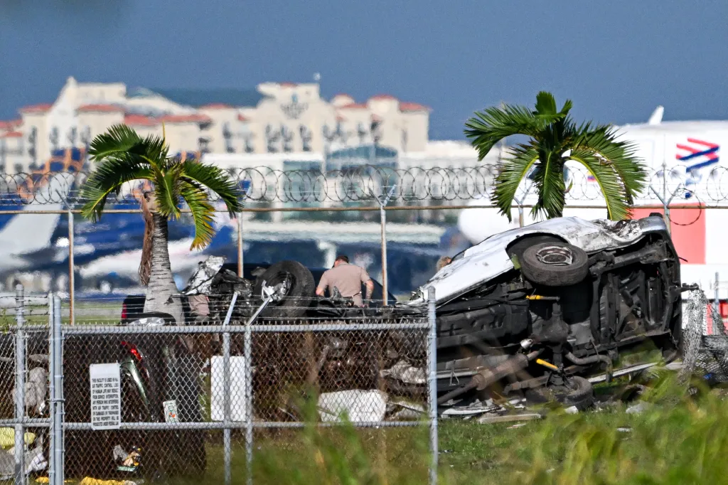 An overturned car and debris from a plane crash at Miami International Airport, with two palm trees and airport buildings in the background.