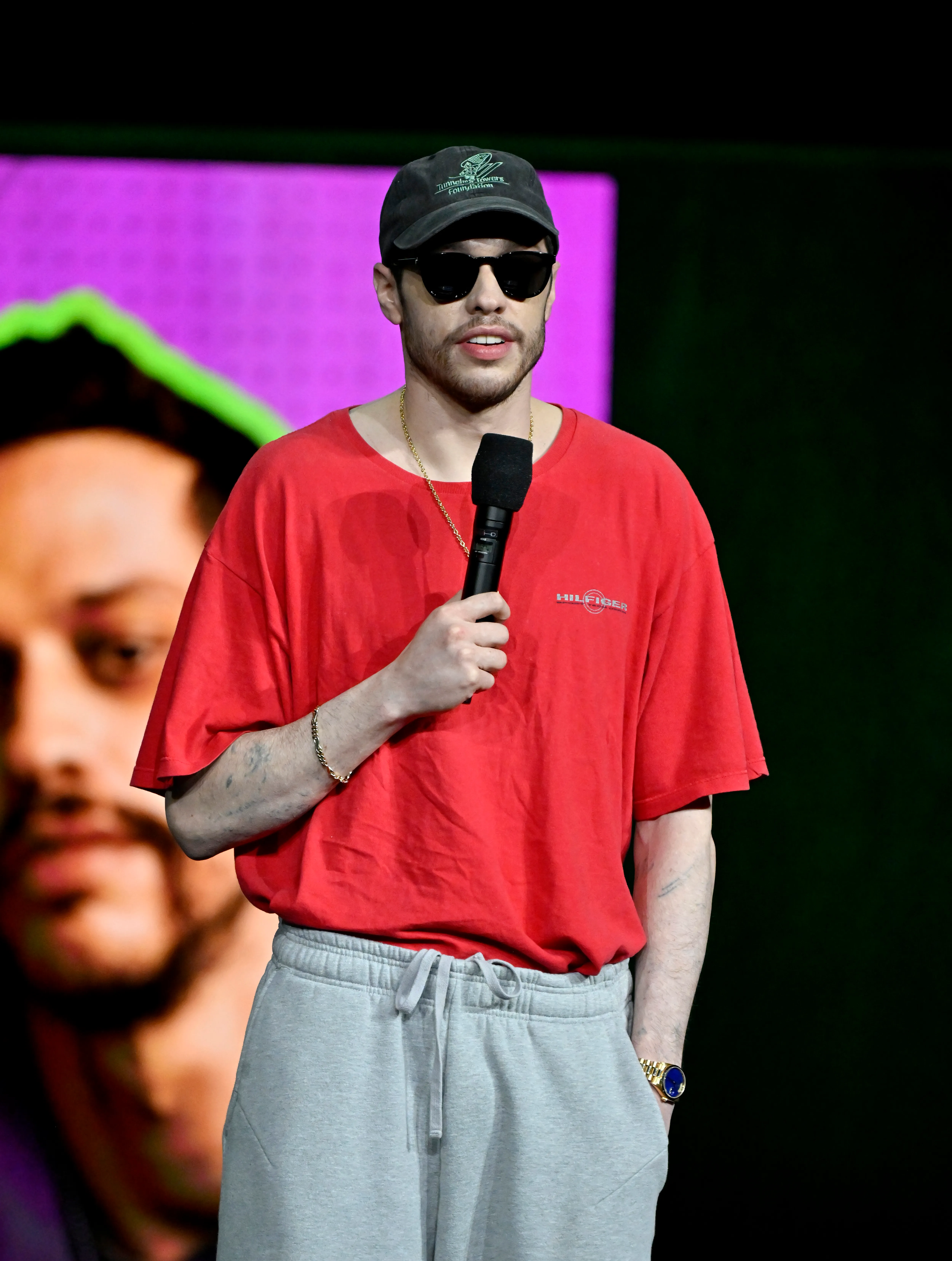 Pete Davidson speaks on stage wearing sunglasses and a red t-shirt.