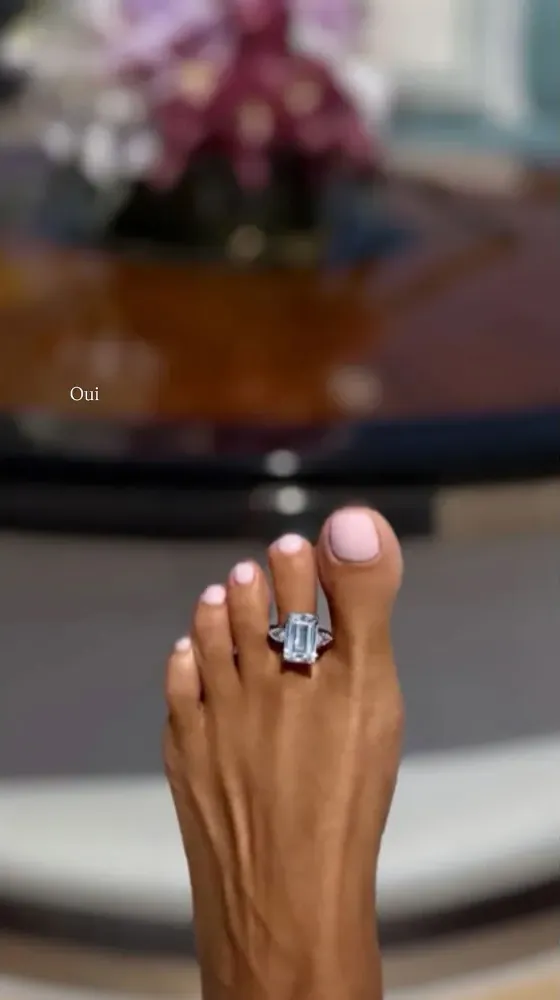 A foot with light pink painted toenails wearing an aquamarine and diamond toe ring on the second toe.