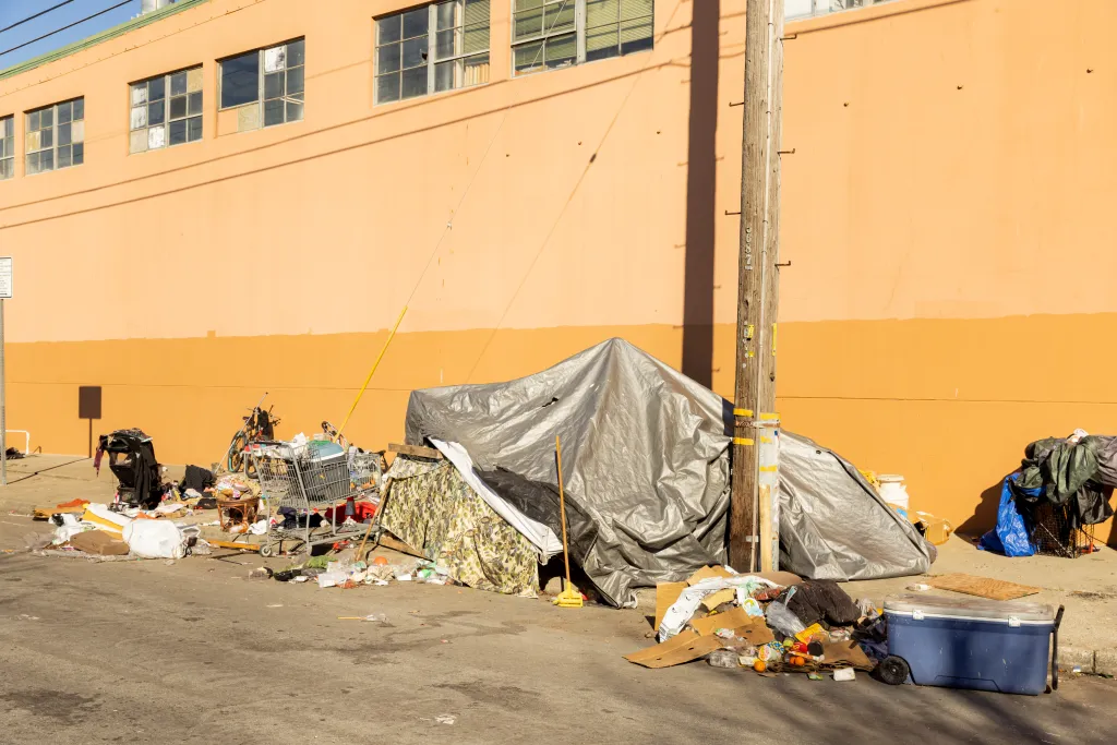 An encampment of tarps and shopping carts filled with belongings, against an orange building in Berkeley, California.