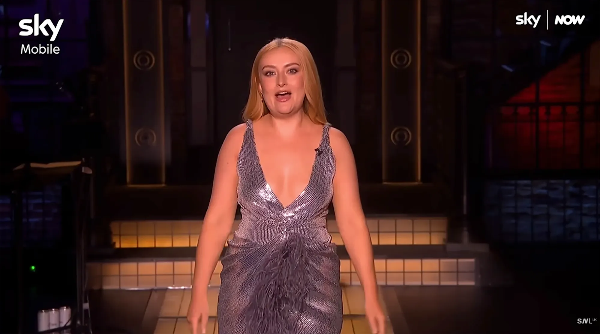 A comedian in a sparkling dress with a deep V-neck and feathery trim on SNL UK.
