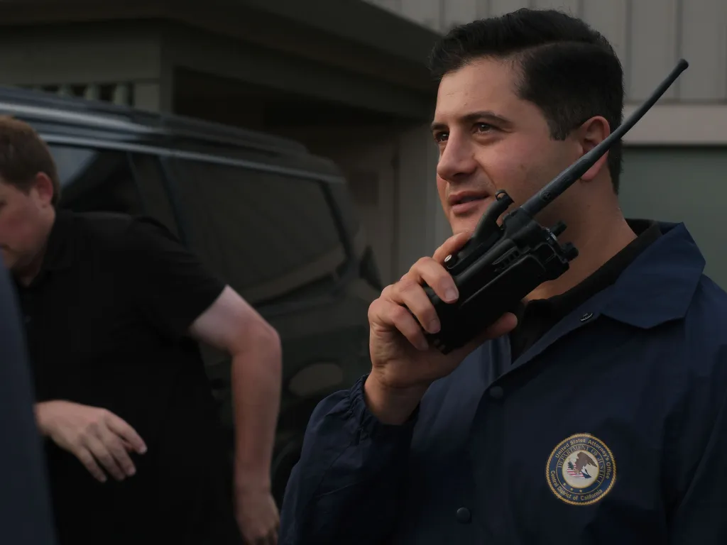An HSI agent wearing a dark blue jacket with a Department of Justice patch on it, holding a radio.