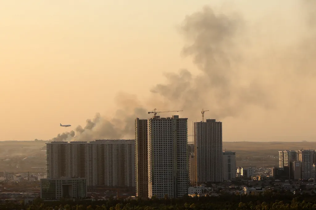 Smoke rises behind city buildings in Erbil, Iraq, with an airplane visible in the sky.