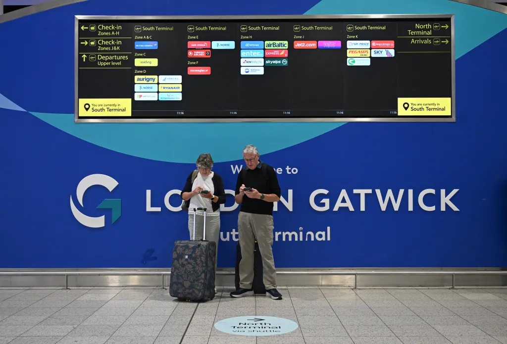 Passengers with luggage consult their phones in the South Terminal of London Gatwick airport, under a flight information display board.