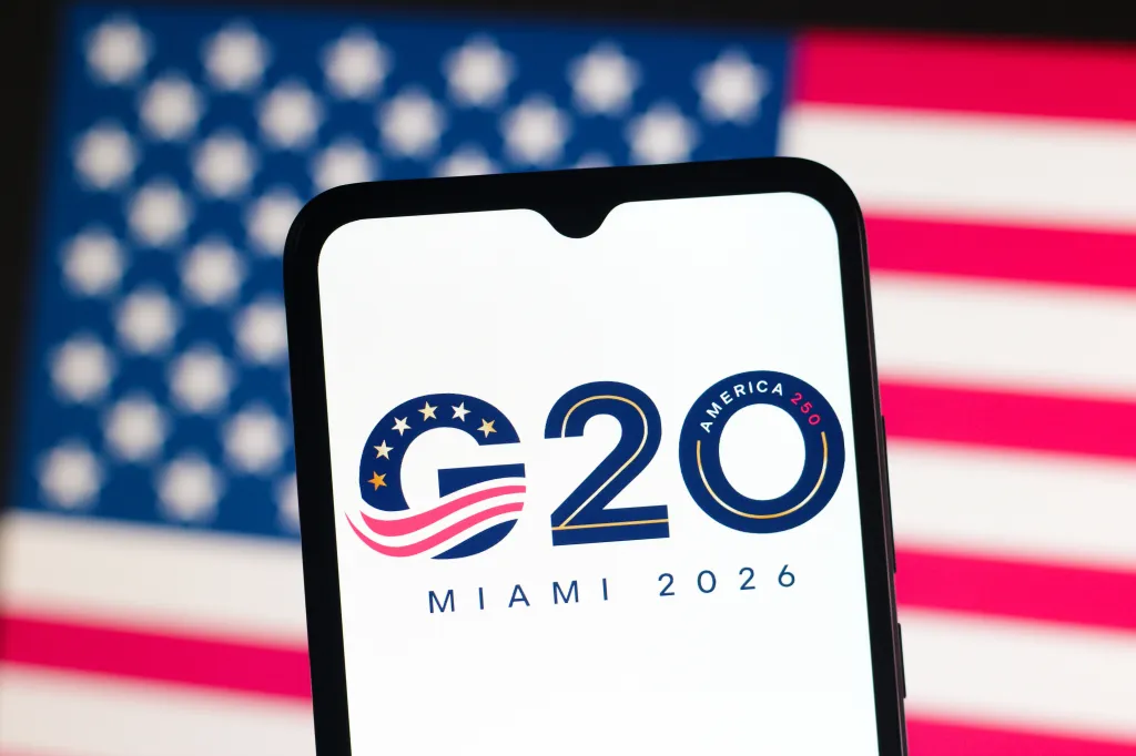 Photo illustration of the 2026 G20 Miami Summit logo on a smartphone with a US flag in the background.