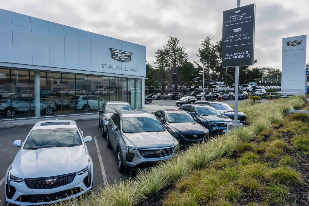 A Cadillac dealership building and sign with several Cadillac cars in the foreground and a parking lot full of cars behind.