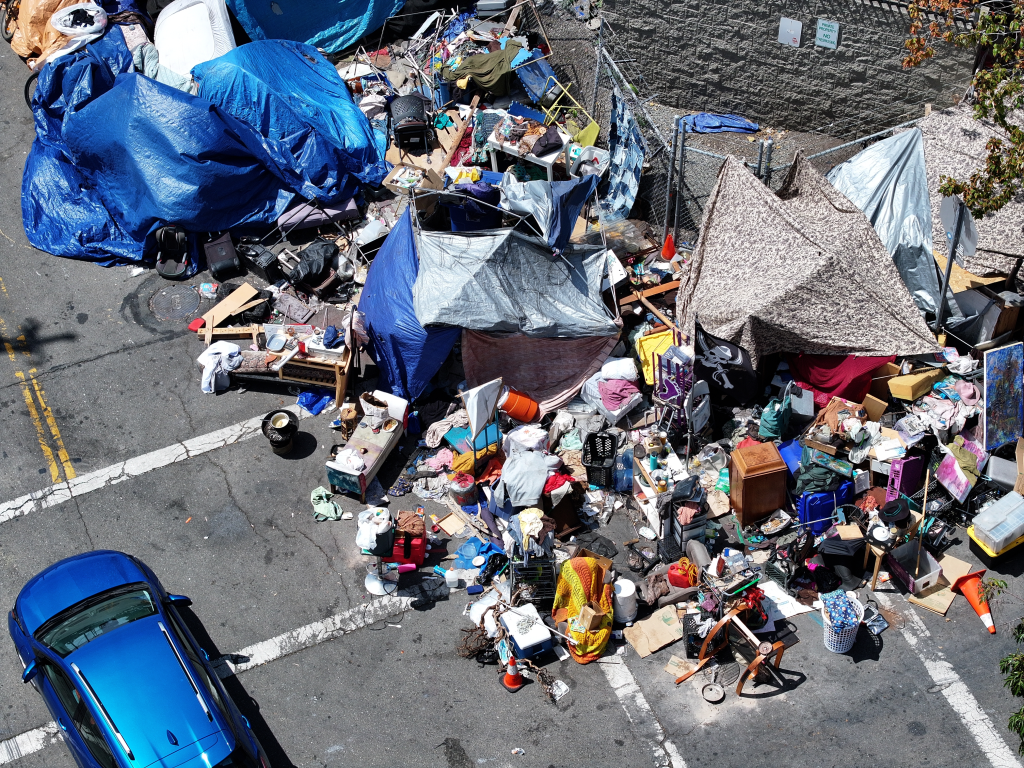Aerial view of a homeless encampment with many tents and tarps, trash, and personal belongings piled high on an asphalt street next to a blue car.