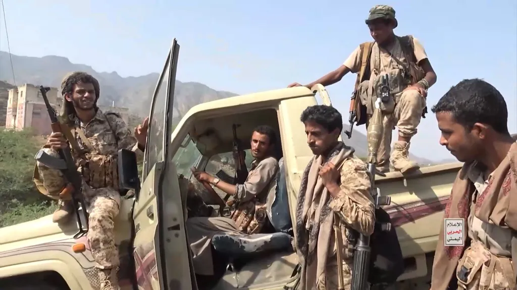 Houthi fighters in camouflage gather around a vehicle, with some carrying rifles and a rocket-propelled grenade launcher.