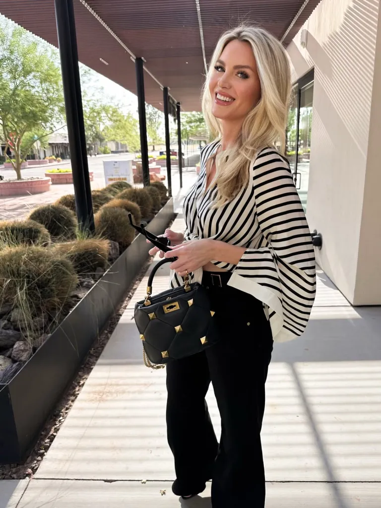 Blonde woman in a striped shirt and black pants holding a black studded purse, smiling while standing outdoors.