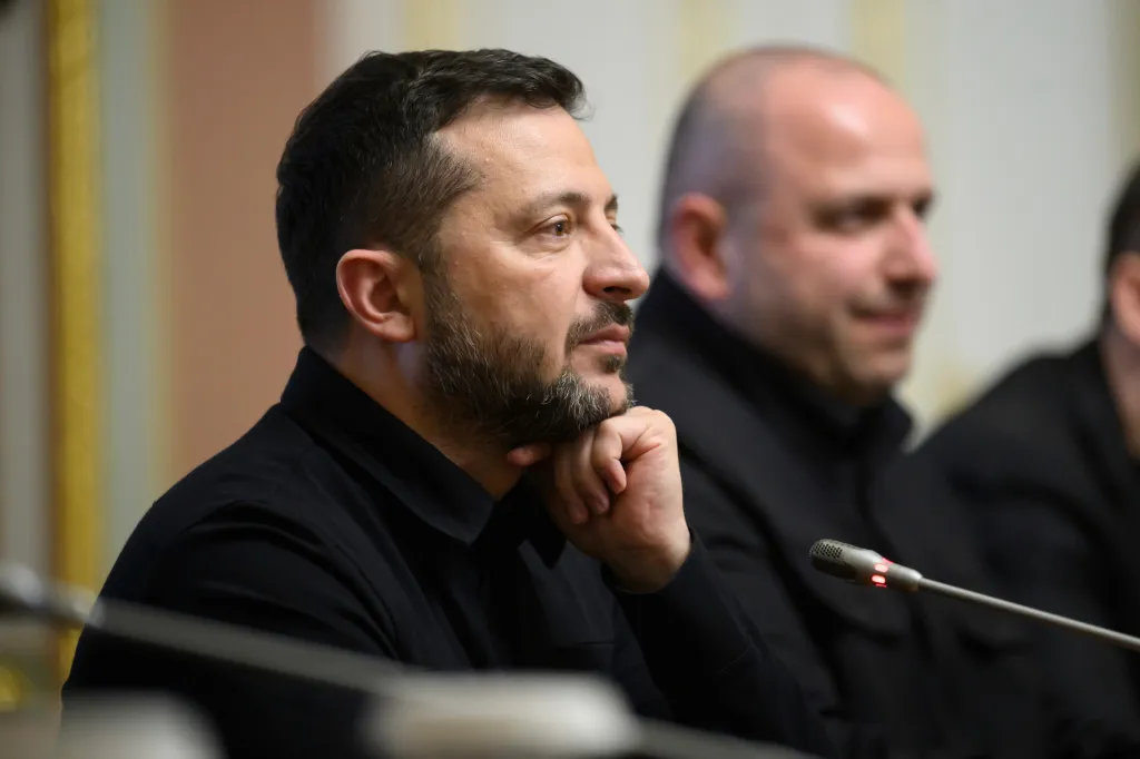 Ukrainian President Volodymyr Zelenskyy, in a black shirt, resting his chin on his fist, looks intently to the right, with a microphone in the foreground and a man blurred in the background.