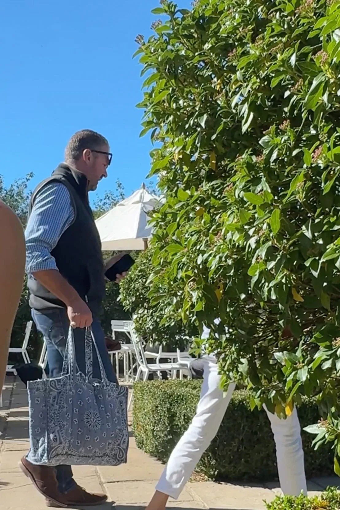 A man in a vest and glasses walks next to a lush green tree, holding a patterned bag.