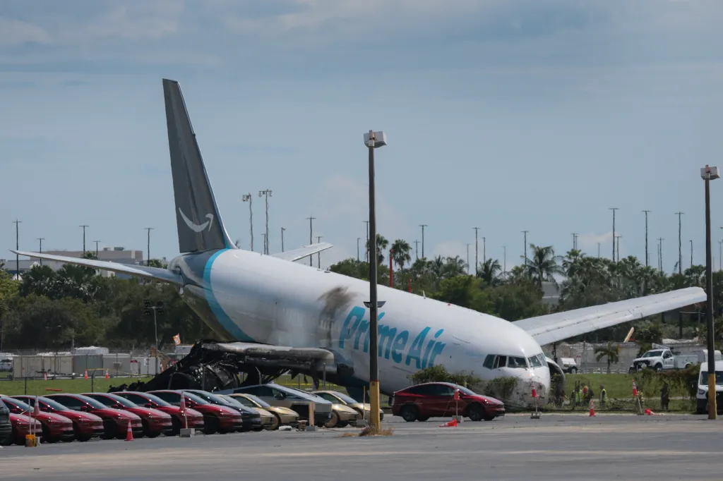 The wreckage of an Amazon Prime Air cargo plane, with a charred engine and its nose resting on the ground, sits on the tarmac near parked cars and palm trees.