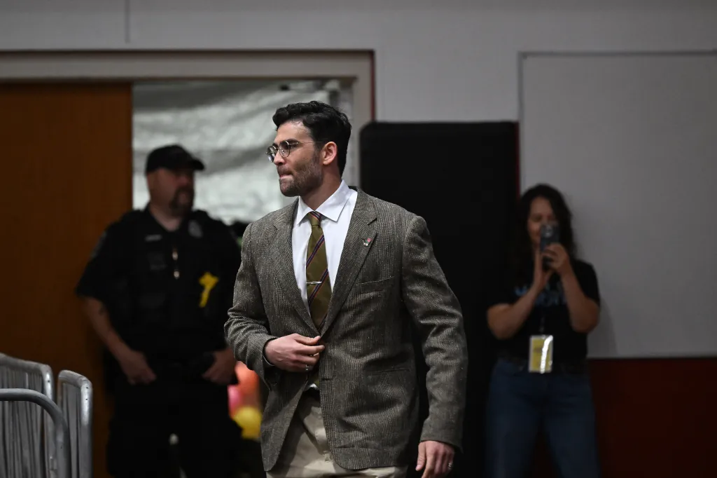 Hasan Piker, wearing a brown suit jacket, white shirt, and striped tie, walks with a hand on his jacket.