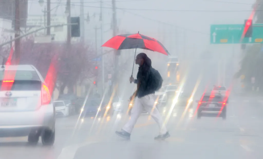 A pedestrian holding a red and black umbrella crosses a wet street in heavy rain with cars and traffic lights visible through the downpour.