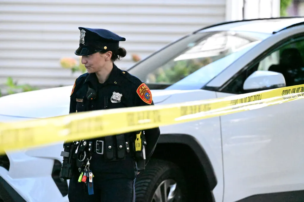 A police officer stands behind yellow crime scene tape in front of a white car.