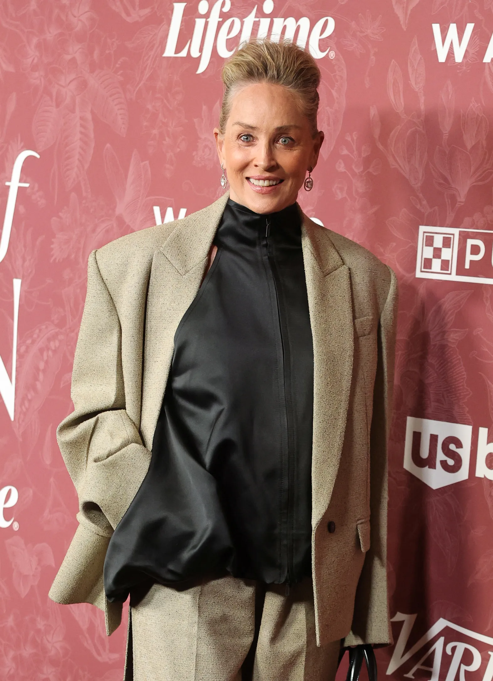 Sharon Stone smiling, wearing a beige suit and black top.