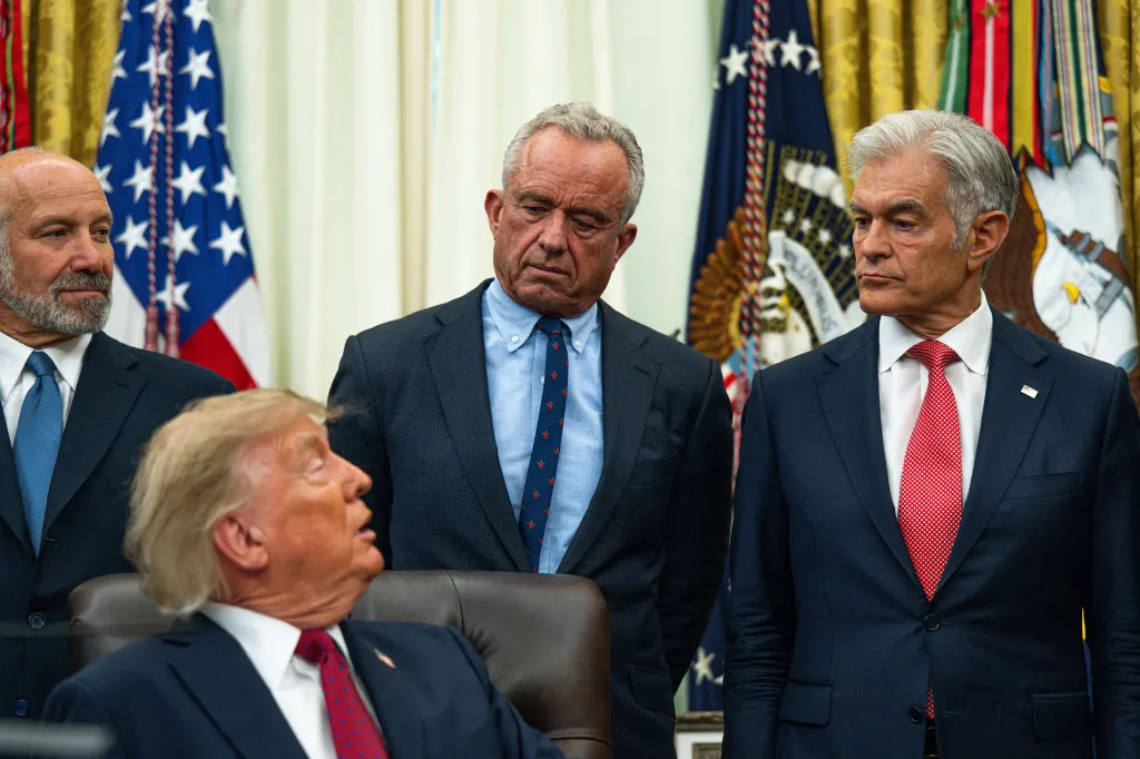 President Trump seated with Robert F. Kennedy Jr. and Mehmet Oz standing behind him in the Oval Office for an announcement on healthcare affordability.