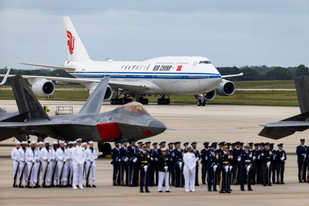 An Air China Boeing 747 aircraft taxis on a runway past military personnel and fighter jets on the tarmac.