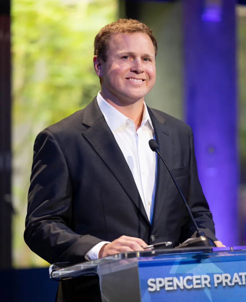 Spencer Pratt smiling at a podium with a microphone.