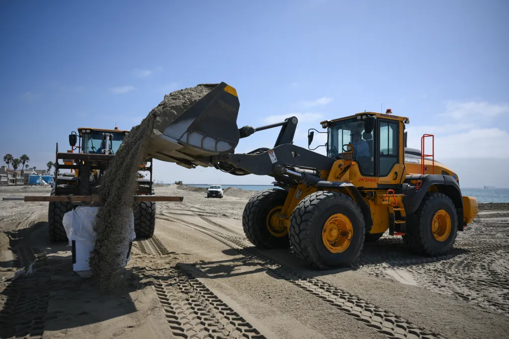 Heavy machinery fills large sandbags on a beach to protect against coastal flooding.